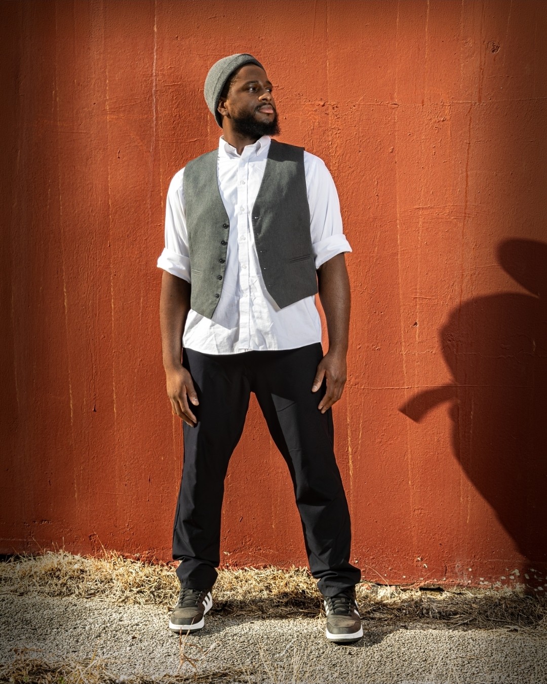 phEEnix standing against a red wall in a grey vest and white button-up shirt